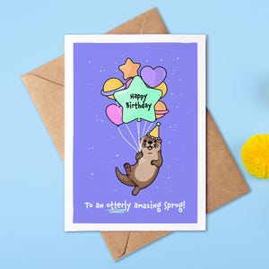 May include: A birthday card with a cartoon otter holding balloons. The card has the text "Happy Birthday" and "To an otterly amazing sprog!" on a purple background. The card is on a brown envelope, with yellow pom-poms.