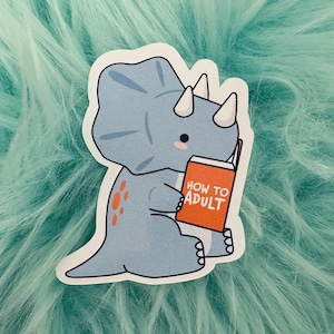 May include: A die-cut sticker featuring a light blue triceratops dinosaur with small white horns and orange spots on its back. The dinosaur is holding an orange book with the white text HOW TO ADULT printed on the front cover in a bold font.