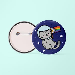 May include: A blue button featuring a grey cat wearing a space helmet and holding a rainbow pride flag in outer space. The cat is floating in a dark blue space with white stars. The reverse of the button is blank.