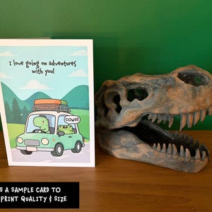 May include: A greeting card featuring a cartoon dinosaur and frog driving a car with the text I love going on adventures with you! and cows!! next to a brown, textured, hollow dinosaur skull replica with sharp teeth.