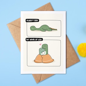 Op de afbeelding: Wenskaart met twee panelen met cartoon dinosaurussen. Het bovenste paneel leest "Always Tired" met een groene dinosaurus die ligt. Het onderste paneel leest "But Never of You" met twee dinosaurussen die elkaar omarmen. De kaart ligt op een bruine envelop.
