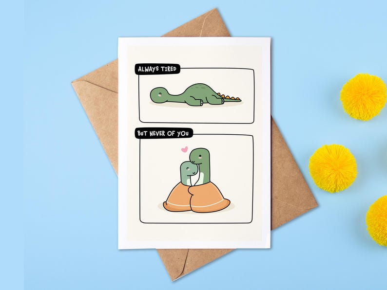 Op de afbeelding: Wenskaart met twee panelen met cartoon dinosaurussen. Het bovenste paneel leest "Always Tired" met een groene dinosaurus die ligt. Het onderste paneel leest "But Never of You" met twee dinosaurussen die elkaar omarmen. De kaart ligt op een bruine envelop.