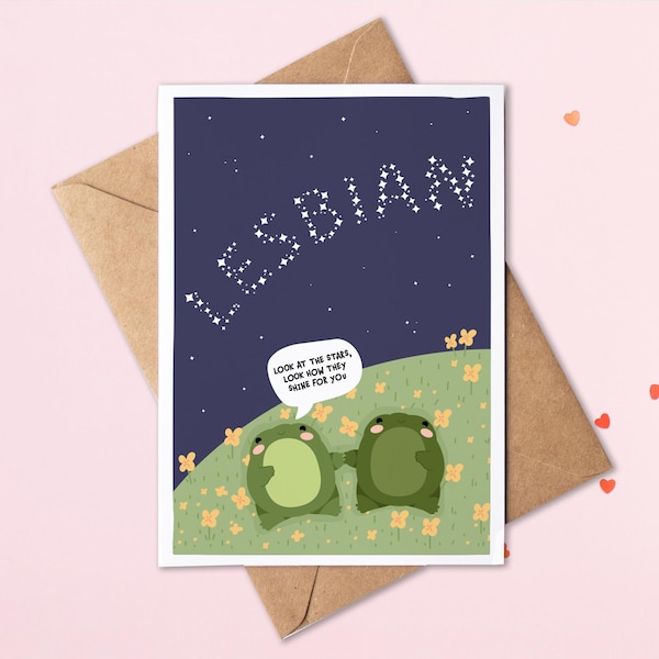 May include: A greeting card featuring two green frogs lying on a grassy hill with yellow flowers under a dark blue starry night sky. The word LESBIAN is written in white stars. A speech bubble says, Look at the stars, look how they shine for you.