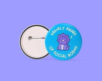 May include: A blue button with a cartoon triceratops and the text "Vaguely Aware of Social Norms." The button is round with a metal pin back. The triceratops is purple and white.