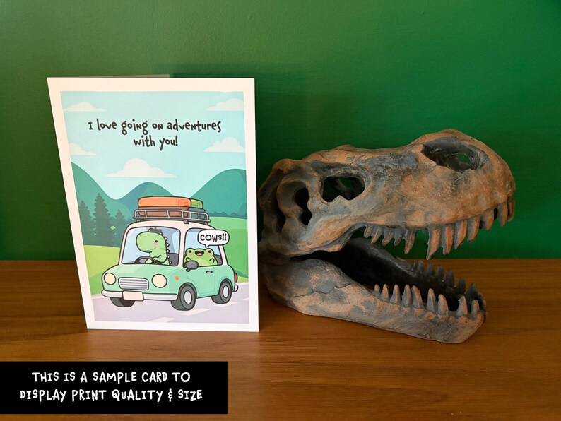 May include: A greeting card featuring a cartoon dinosaur and frog driving a car with the text I love going on adventures with you! and cows!! next to a brown, textured, hollow dinosaur skull replica with sharp teeth.