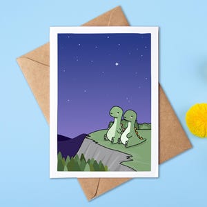 Puede incluir: Una tarjeta de felicitación rectangular con una ilustración de dibujos animados de dos dinosaurios verdes sentados en el borde de un acantilado por la noche. La escena muestra un cielo azul oscuro estrellado sobre un paisaje con colinas verdes, árboles y un sobre marrón.