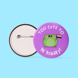 May include: A purple button with the text "TOO CUTE TO BE BINARY!" and a cartoon frog holding a non-binary pride flag. The button is next to a button back with a pin. The background is light blue.
