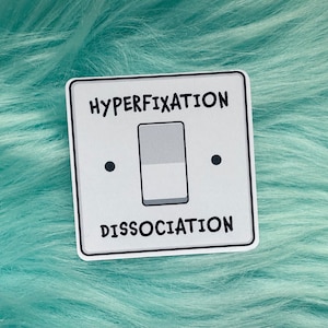 May include: A square sticker featuring a light grey light switch plate design. The word HYPERFIXATION is printed in black above the switch, and the word DISSOCIATION is printed in black below the switch. The switch itself is grey and white.