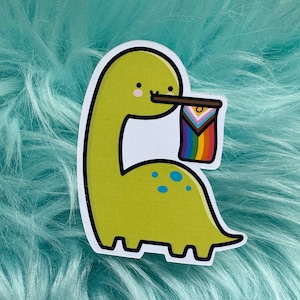 May include: A green cartoon dinosaur holding a pride flag with a yellow, white, pink, blue, green, turquoise, and purple design. The dinosaur has a small smile and a single blue eye.