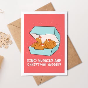 Op de afbeelding: Een wenskaart met een koraalrode achtergrond met een lichtblauwe afhaaldoos gevuld met bruine dinosaurusvormige kipnuggets die kleine rode kerstmutsen dragen. De tekst DINO NUGGIES AND CHRISTMAS HUGGIES is in het wit gedrukt.