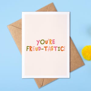 May include: A rectangular greeting card with a light pink background featuring the colorful text "YOU'RE FREUD-TASTIC!" in a playful, multi-colored font. The card is placed on top of a brown kraft paper envelope.