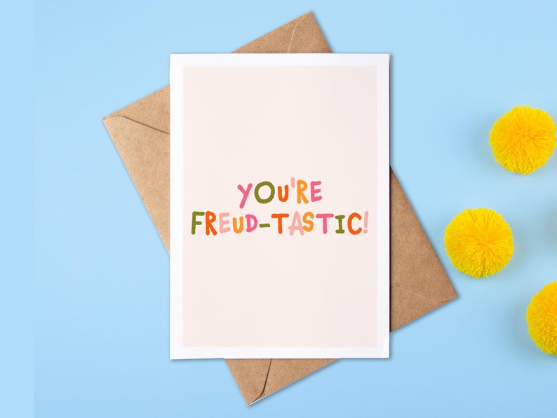 May include: A rectangular greeting card with a light pink background featuring the colorful text "YOU'RE FREUD-TASTIC!" in a playful, multi-colored font. The card is placed on top of a brown kraft paper envelope.