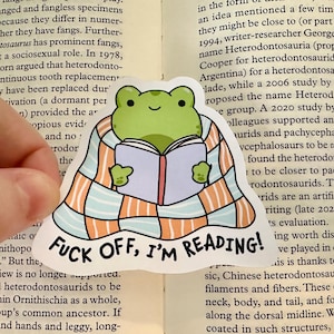 Op de afbeelding: Een gestanste sticker met een groene kikker gewikkeld in een lichtblauwe en oranje geruite deken terwijl deze een open boek leest. De tekst "FUCK OFF, I'M READING!" staat in zwarte hoofdletters gedrukt aan de onderkant van de sticker.