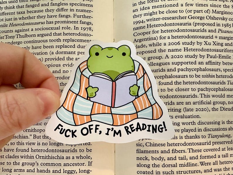 Op de afbeelding: Een gestanste sticker met een groene kikker gewikkeld in een lichtblauwe en oranje geruite deken terwijl deze een open boek leest. De tekst "FUCK OFF, I'M READING!" staat in zwarte hoofdletters gedrukt aan de onderkant van de sticker.
