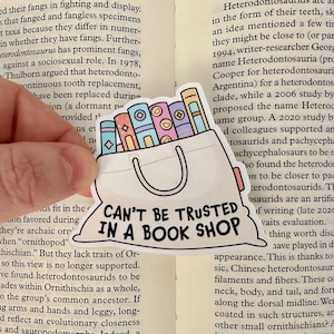 Puede incluir: Una pegatina blanca con un contorno negro de una bolsa llena de libros coloridos. El texto "Can't be trusted in a book shop" está escrito en la bolsa.