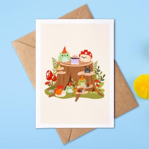 May include: A greeting card with a whimsical illustration of a tree stump with cartoon frogs, mushrooms, and a cake. The card is on a brown envelope, with yellow pom-poms on a blue background.