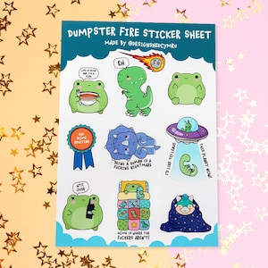 May include: A rectangular sticker sheet titled DUMPSTER FIRE STICKER SHEET MADE BY @DESIGNSHEDCYMRU featuring various cartoon illustrations including frogs, a dinosaur, a blue ribbon, a UFO, and text captions like LIFE IS SOUP AND I'M A FORK.