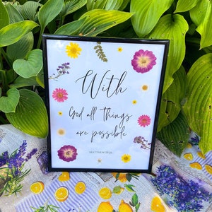 May include: A framed print with a white background and black frame. The print features a floral design with pressed flowers and the text "With God all things are possible. Matthew 19:26".