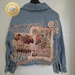Puede incluir: Una chaqueta vaquera azul claro con un panel trasero decorado con un collage de encaje y un parche de tela estampado. El parche muestra una escena de un anfiteatro clásico junto a un grupo de casas coloridas y árboles de pino.
