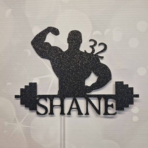 May include: Black glitter cake topper featuring a silhouette of a weightlifter lifting a barbell. The weightlifter has one arm raised and the other arm bent at the elbow. The barbell has weight plates on each end. The number 32 is on the weightlifter's right side. The name "SHANE" is written below the barbell.