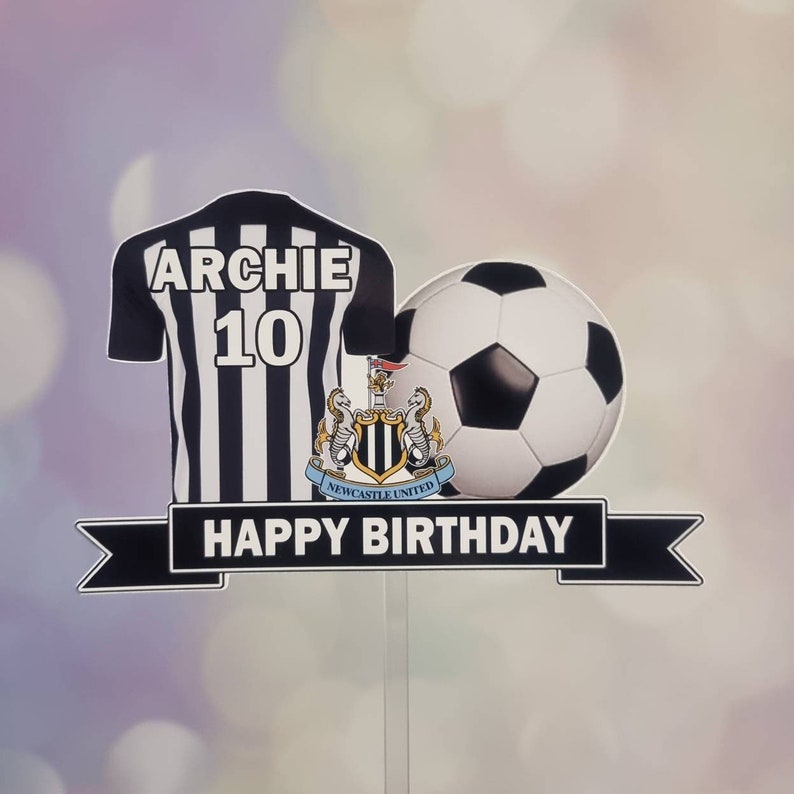 May include: Black and white football shirt with the name "Archie" and the number "10" on the back. A black and white football and a Newcastle United crest are on top of the shirt. A black ribbon with the text "Happy Birthday" is below the shirt.