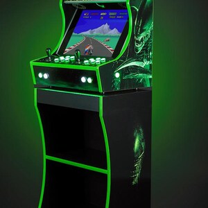 May include: A black and green arcade cabinet with a green neon outline. The cabinet has a screen displaying a video game with the text "ALIENS" on the top. The cabinet has a stand with two shelves.