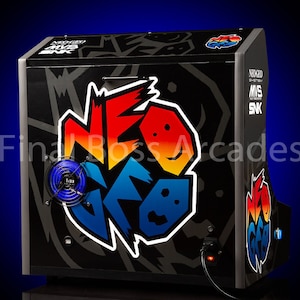 May include: A black arcade cabinet with a colourful "NEO GEO" logo on the front. The cabinet has a blue fan on the left side and a power cord plugged in on the right side. The text "NEO GEO MVS SNK" is printed on the right side of the cabinet.