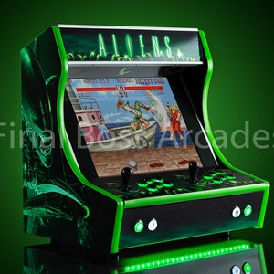 May include: A black and green arcade cabinet with a green neon light on the bottom. The cabinet has a screen showing a video game with the text "ALIENS" on the top.