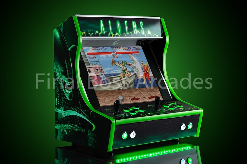 May include: A black and green arcade cabinet with a green neon light on the bottom. The cabinet has a screen showing a video game with the text "ALIENS" on the top.