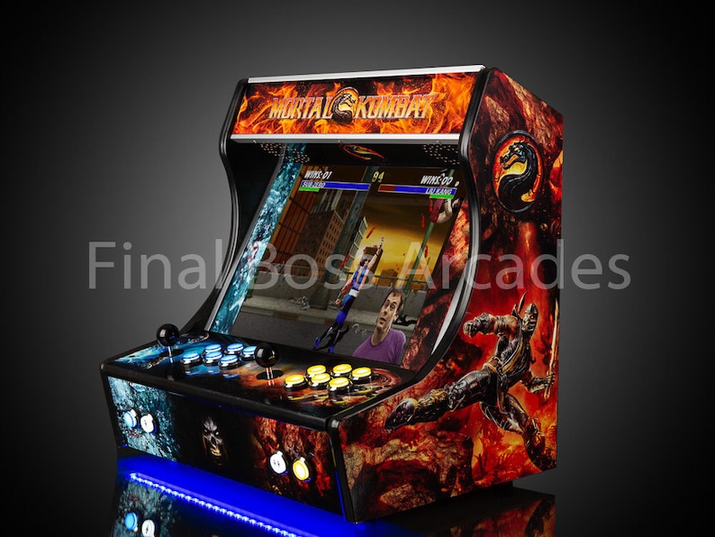 May include: A black arcade cabinet with a Mortal Kombat theme. The cabinet features a red and orange flame design with a black and silver Scorpion character on the side. The cabinet has a blue light underneath it.