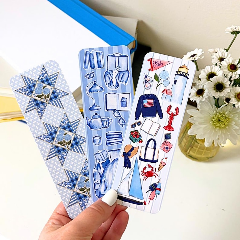 May include: Three decorative bookmarks with blue and white designs. One features a star pattern, another has illustrations of books and a teapot, and the third depicts coastal elements like a sailboat and lobster.