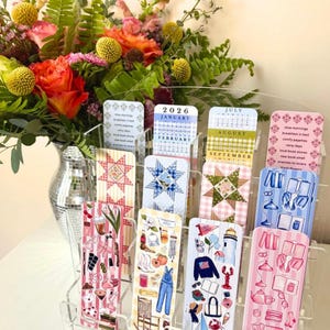 May include: A clear acrylic display holding decorative bookmarks and a small calendar for 2026. The bookmarks feature various designs, including quilt patterns and illustrations of everyday objects. A vase of colorful flowers is in the background.