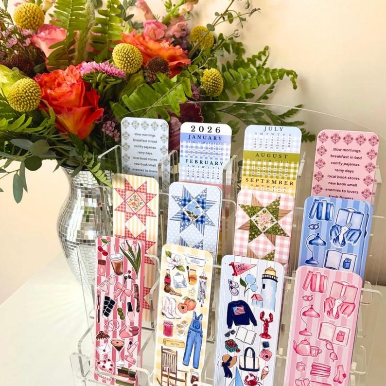 May include: A clear acrylic display holding decorative bookmarks and a small calendar for 2026. The bookmarks feature various designs, including quilt patterns and illustrations of everyday objects. A vase of colorful flowers is in the background.