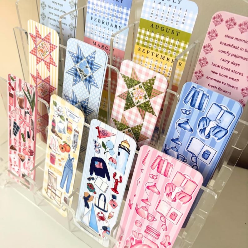 May include: A clear acrylic display holding several decorative bookmarks. The bookmarks feature various designs, including floral patterns, calendar months, and illustrated scenes. Some bookmarks have text like "AUGUST" and "EAST COAST".