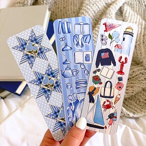 May include: Three decorative bookmarks with blue and white designs. One features a star pattern with floral accents, another has book-themed illustrations, and the third showcases East Coast imagery, including a sailboat and lobster.
