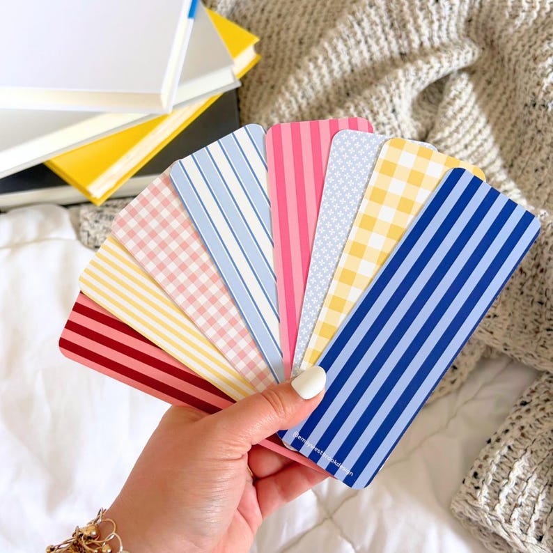 May include: A hand holds a fan of patterned bookmarks. The bookmarks feature various designs, including stripes, gingham, and floral patterns in shades of red, pink, yellow, and blue. A stack of books with yellow and white covers is in the background.