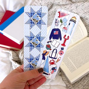 May include: Two decorative bookmarks. One features a blue and white star pattern with floral accents. The other has a nautical theme with illustrations of a lighthouse, lobster, sailboat, and a sweater with an American flag.