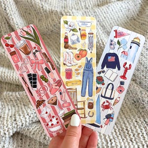 May include: Three illustrated bookmarks with different themes. One features cherries, a drink, and a cake. Another has overalls, peaches, and cowboy boots. The third shows a lighthouse, lobster, and a sweater with an American flag.