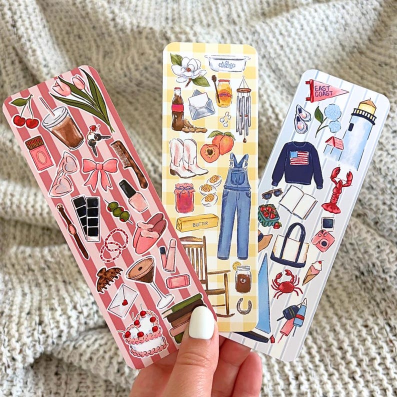 May include: Three illustrated bookmarks with different themes. One features cherries, a drink, and a cake. Another has overalls, peaches, and cowboy boots. The third shows a lighthouse, lobster, and a sweater with an American flag.