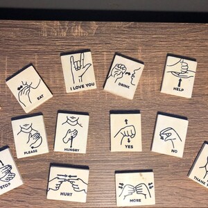 May include: A set of 15 wooden tiles with black illustrations of sign language hand gestures and the corresponding words printed below each image. The words include "Sorry", "Eat", "I Love You", "Drink", "Help", "Bathroom", "Please", "Hungry", "Yes", "No", "Stop", "Hurt", "More", and "Thank You".