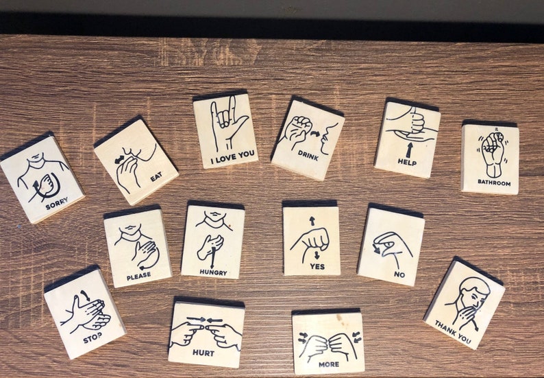 May include: A set of 15 wooden tiles with black illustrations of sign language hand gestures and the corresponding words printed below each image. The words include "Sorry", "Eat", "I Love You", "Drink", "Help", "Bathroom", "Please", "Hungry", "Yes", "No", "Stop", "Hurt", "More", and "Thank You".