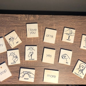 Può includere: Un set di 14 carte di linguaggio dei segni in legno con illustrazioni e testo neri. Le carte includono segni per "mi dispiace", "mangiare", "ti amo", "bere", "aiutare", "bagno", "fermare", "per favore", "affamato", "sì", "no", "male", "di più", e "grazie".