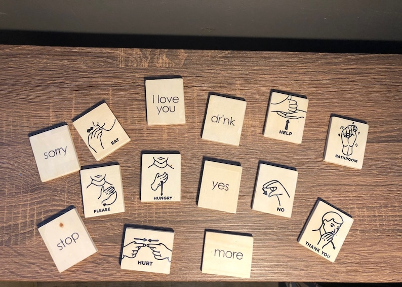 May include: A set of 14 wooden sign language cards with black illustrations and text. The cards include signs for "sorry", "eat", "I love you", "drink", "help", "bathroom", "stop", "please", "hungry", "yes", "no", "hurt", "more", and "thank you".