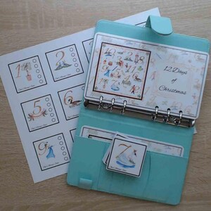 May include: A turquoise planner with a white insert featuring the 12 Days of Christmas. The insert has a colorful illustration of the 12 days of Christmas with numbers 1-12. The planner has a ring binder system and is open to reveal the insert. There are also 12 individual cards with numbers 1-12, each with a corresponding illustration.
