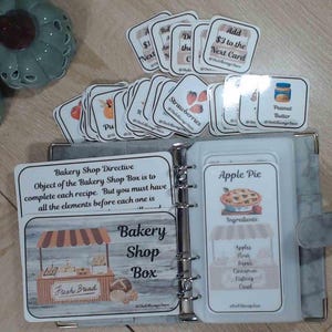 Puede incluir: Un archivador abierto con tarjetas de recetas y tarjetas de ingredientes. Las tarjetas presentan ilustraciones de frutas, productos horneados y texto. El archivador está abierto para mostrar las tarjetas, con una cubierta gris.