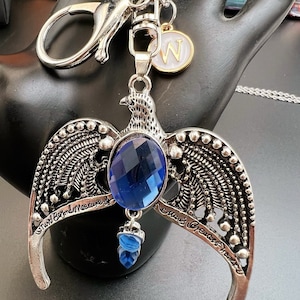 May include: Silver-toned keychain featuring a stylized eagle pendant with a large, faceted blue gemstone and a smaller blue teardrop-shaped gem. The pendant has engraved text and is attached to a keyring with a lobster clasp and a charm with the letter "W".