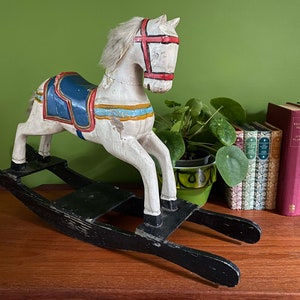 May include: A white wooden rocking horse with blue and red painted details. The horse has a saddle and a bridle. The rocking horse is on a black wooden base.