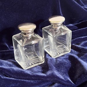 May include: Two clear glass bottles with square bases and rounded silver-toned metal caps. Each bottle features a starburst pattern etched into the bottom and faceted edges along the sides, creating a geometric appearance for these containers.