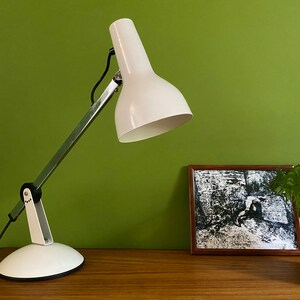 May include: A white desk lamp with a silver arm and a white base. The lamp is on a wooden table with a framed black and white print and a potted plant.