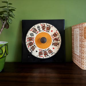 May include: A black square frame containing a round, white card game wheel. The wheel has a yellow centre and is surrounded by playing cards with red and black suits. The frame is on a wooden shelf with a green plant in a green pot to the left and a row of four books to the right.